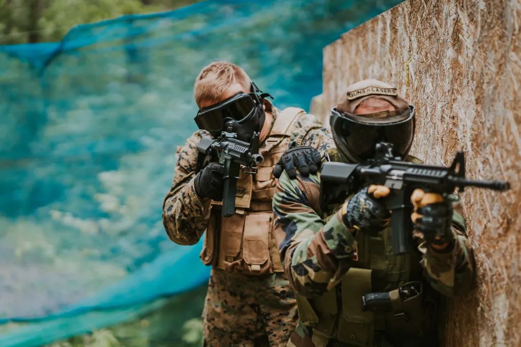 Divi airsofta spēlētāji ekipējumā aiz aizsega āra laukumā